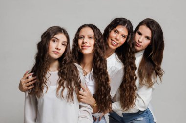 Uzun saçlı dört genç kadın birlikte poz veriyor, düz gri bir arka plana gülümsüyor, günlük beyaz ve kot giysiler giyiyor, cazibe, arkadaşlık ve birliktelik yayıyor..