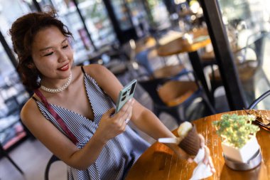 Kafede gülümseyen genç bir kadın ferahlatıcı bir dondurma külahının tadını çıkarırken akıllı telefon kullanıyor. Ilık bir ışık ve rahat bir atmosferle rahat bir an..