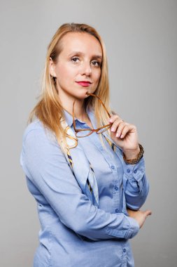 Bir iş kadını soğukkanlı bir ifadeyle ayakta duruyor, bir elinde gözlüğünü hafifçe tutuyor. Açık mavi bir gömlek giyiyor. Tarafsız bir arkaplanda özgüven ve profesyonellik yayıyor..