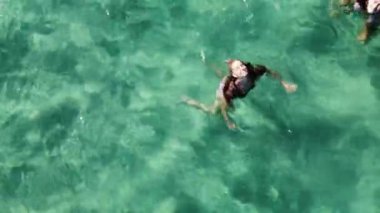 İki genç kadın kristal berrak turkuaz suda yüzüyor. Elleriyle kalp şekilleri yapıp kameraya gülümsüyorlar..