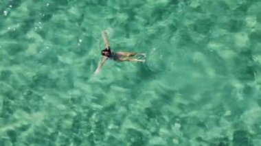 Yüzen, yüzen ve elleri turkuazda, şeffaf deniz suyunda kalp yapan bir kadın. Havadan yukarıdan aşağıya görünüm.