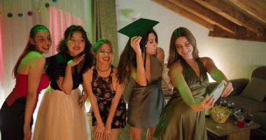 Arkadaşlar mezuniyeti ev partisinde kutluyor. Şık kızlar özçekim için poz veriyor, ellerinde mezuniyet şapkası ve diploması var. Şenlik havası, zarif elbiseler, neşe ve birliktelik..