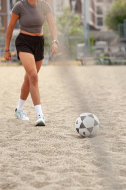 Kırpılmış bluzlu ve şortlu bir sporcu, kumlu bir sahada desenli bir topu tekmeliyor..