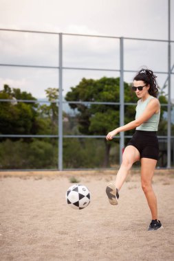 Nane rengi şortlu bir sporcu açık gökyüzünün altındaki kumlu bir sahada futbol topunu tekmeliyor..