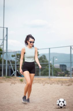 Hafif kolsuz tişörtlü ve siyah şortlu atletik kadın tozlu bir sahada futbol topunu tekmeliyor..