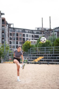 Açık havada antrenman yapan atletik bir kadın, arka planda şehir binaları olan kumlu bir sahada futbol topu tekmeliyor..