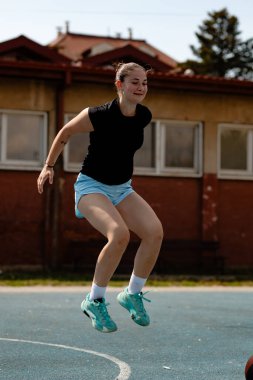 Bir basketbol oyuncusu hava yoluyla antrenman sırasında açık bir sahada atlıyor. Parlak güneş ışığı, ağa nişan aldığı ve yeteneklerini geliştirdiği için odaklanmış yüz ifadesini vurgular..