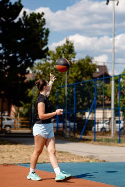 Bir basketbol oyuncusu dışarıda bir sahada duruyor, top tutuş ve atış antrenmanına odaklanıyor. Güneş parlıyor, ağaçlar ve kentsel elementlerle dolu canlı bir atmosfer yaratıyor..