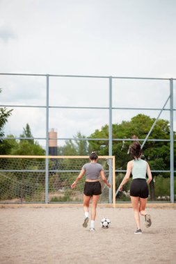 Toprak bir sahada futbol oynayan iki kadın, sıradan bir antrenman sahnesinde topun peşinden koşuyor..