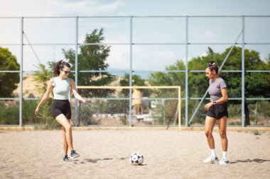 İki kadın kumlu bir sahada futbol antrenmanı yapıyor. Onlar bir spor kompleksinde becerileri üzerinde çalışırken topa odaklanırlar. Hava güneşli ve açık hava aktivitesi için güzel bir gün..