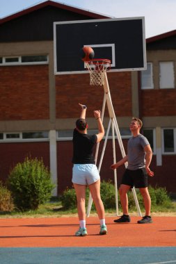İki oyuncu açık hava sahasında sıradan bir basketbol antrenmanına katılıyor. Bir oyuncu basketi atarken diğeri güneşli günün tadını çıkarıyor..