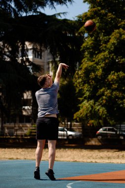 Genç bir erkek oyuncu, parlak güneş ışığı altında iyi korunan bir açık hava sahasına basketbolu elinden bırakırken dikkatle odaklanıyor. Alanı çevreleyen uzun ağaçlar.