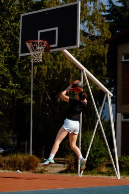 Bir basketbol oyuncusu havalandı ve topu açık hava sahasında potaya fırlatmaya hazırlanıyor. Ağaçlar ve binalar enerjik atmosferi geliştirmek için canlı bir zemin oluşturur..