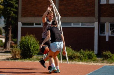 İki genç basketbolcu yeteneklerini parlak bir öğleden sonra, yeşilliklerle çevrili bir açık hava basketbol sahasında sergiliyorlar ve savunuyorlar..