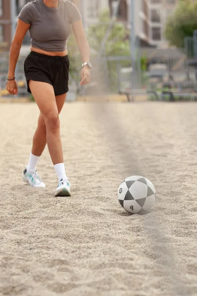 Kırpılmış bluzlu ve şortlu bir sporcu, kumlu bir sahada desenli bir topu tekmeliyor..