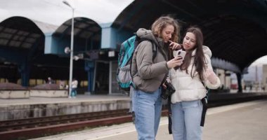 Sırt çantalı ve kameralı iki genç kadın tren istasyonunda telefonlarındaki resimlere bakıp gülüyorlar..