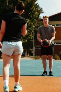 İki basketbolcu açık hava sahasında antrenman yapıyor. Bir oyuncu pas vermeye hazırlanırken diğeri topu almaya hazır. Gün aydınlık ve açık.