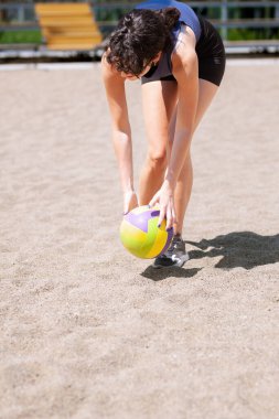 Bir kadın güneşli bir günde kumdan parlak renkli bir voleybol topu almak için eğilir. Voleybol sahası açık, eğlenceli bir açık hava aktivitesini yansıtıyor..