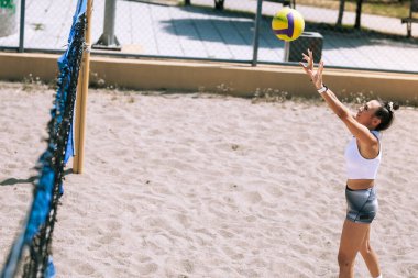 Beyaz bluzlu ve gri şortlu bir oyuncu kumlu bir sahada voleybol oynamaya hazırlanıyor. Güneş ışıl ışıl parlıyor ve atmosfer canlı..