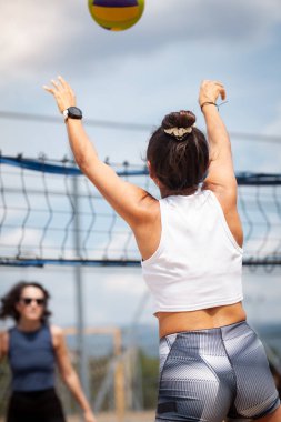 İki oyuncu açık havada dostça bir voleybol maçı yapıyor. Bir oyuncu servis yapmaya hazırlanırken diğeri dikkatle izliyor. Sahnede kumlu bir kort, ağlar ve güneşli bir hava yer alıyor..