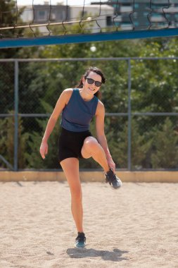 Parktaki bir voleybol sahasında duran bir kadın bacağını esnetiyor. Sıcak güneşin tadını çıkarıyor ve arkadaşlarıyla bir oyuna hazırlanıyor..