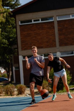 Oyuncular açık hava sahasında zorlu bir basketbol maçının keyfini çıkarırken, açık mavi gökyüzünün altında canlı bir ortamda yeteneklerini sergiliyorlar..