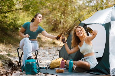 İki arkadaş ormanda çadırlarının yanında oturuyorlar. Biri selfie çekerken diğeri içkiyle tezahürat yapıyor. Yeşillik onları çevrelemiş, neşeli bir açık hava ortamı yaratmış..