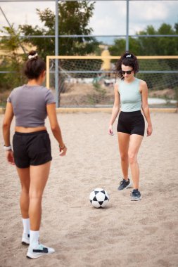 İki kadın tozlu bir sahada futbol oynuyor, biri naneli tişörtlü, diğeri gri şortlu, topa odaklanmış..