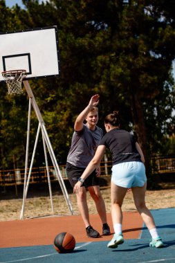 Açık gökyüzünün altında, iki basketbolcu açık hava sahasında antrenman yapıyor. Çemberin yanındaki oyuncu savunmasını yaparken, diğeri de topu sürmeye hazırlanıyor. Yetenek ve takım çalışması sergiliyor..