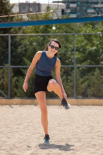 Parktaki bir voleybol sahasında duran bir kadın bacağını esnetiyor. Sıcak güneşin tadını çıkarıyor ve arkadaşlarıyla bir oyuna hazırlanıyor..
