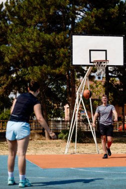 Güneşli bir sahada iki basketbol oyuncusu antrenman yapıyor. Bir oyuncu pas verirken diğeri potaya doğru atış yapmaya hazırlanıyor, takım çalışması ve yetenek sergiliyor..