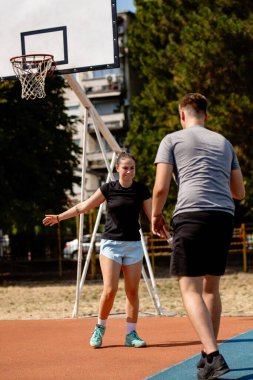 İki basketbolcu, güneşli bir günde şehir parkındaki bir sahada antrenman yapıyor. Bir oyuncu hareket ederken diğeri de karşılık vermeye hazırlanıyor..