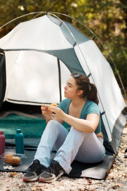 Ormanlık bir alanda çadırın dışında oturan bir kadın gündelik kıyafetlerle atıştırmanın tadını çıkarıyor. Güneş ışığı ağaçların arasından süzülüp, dışarıda huzurlu bir atmosfer yaratıyor..