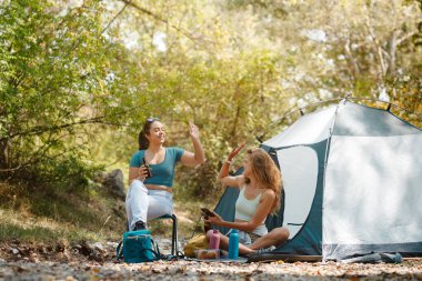 Arkadaşlar çadırlarının dışında ağaçlarla çevrili olarak rahatça otururlar. Sohbete dalıyorlar, içkilerini yudumluyorlar ve güneşin sıcaklığının tadını çıkarıyorlar..