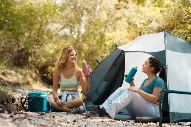 İki genç kadın yemyeşil bir alanda çadırlarının dışında rahatça oturuyorlar. Gülüşlerini ve içkilerini paylaşıyorlar, doğada birlikte geçirdikleri zamanın tadını çıkarıyorlar. Güneş ışığı ağaçların arasından süzülür..