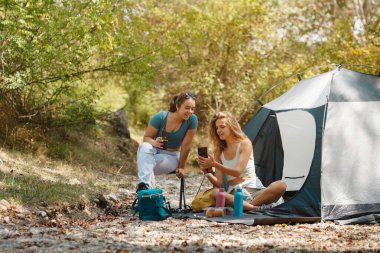 İki arkadaş huzurlu bir ormanda çadırlarının yanında oturuyorlar. Güneşli bir günde doğayla iç içe gülümseyip telefona bakıyorlar. Atıştırmalıklar ve içecekler yakında..