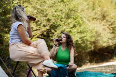 İki kadın doğal ortamda kamp yaparken içki içip sohbet ediyorlar. Ağaçlarla çevrili bir battaniyenin üzerinde otururlar. Açık havanın ve birlikte geçirdikleri zamanın tadını çıkarırlar..