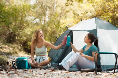 Bir kamp alanındaki arkadaşlar, ağaçlar ve doğa tarafından çevrelenmiş bir şekilde, sıcak güneşin altında, yerde oturmuş, gülümsüyor ve içecek şişelerini kaldırıyorlar..
