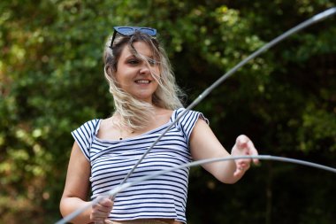 Bir kız güneşli bir günde kamp alanında iplerle ustalıkla oynayarak eğleniyor. Saçları rüzgarda uçuşuyor. Gülümserken, açık hava aktivitelerinin neşesini gösteriyor..