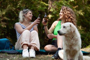 Ormandaki bir paspasın üzerinde oturan iki kadın, yakınlarda bir köpek dinlenirken gülümseyerek içkilerini tokuşturuyorlar. Onların kaygısız varlığı güneşli bir öğleden sonra kamp yapmanın neşesini yakalar..