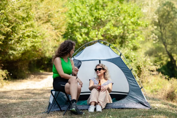 İki kadın çadırlarının dışında oturmuş, rahat bir kamp deneyiminin tadını çıkarıyorlar. Etrafları yemyeşil ve güneş ışığıyla çevrili, hikayelerini ve kahkahalarını doğalarında sükunetle paylaşıyorlar..