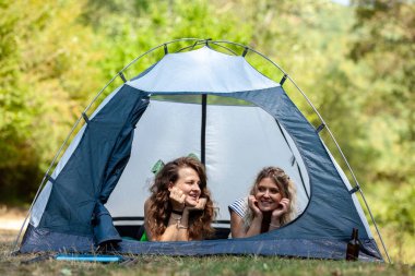 Kadınlar çadırın içinde rahatça uzanır, doğanın ortasında gülümser ve sohbet ederler. Sahne, güneşli bir günde verimli ve ağaçlık bir bölgede neşeli bir kamp deneyimi yakalar..