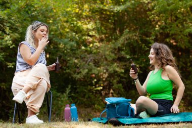 İki kadın ormanda bir piknik battaniyesinde oturmuş, soğuk içeceklerin tadını çıkarıyor ve ağaçlar ve doğa güzellikleriyle çevrili sıcak güneşin altında dinleniyorlar..