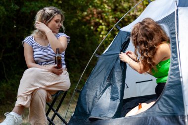 İki kadın bir kamp deneyimi yaşarken, biri çadırın önünde içki içerken, diğeri çadırın girişini ayarlıyor. Çevreleyen doğa canlı ve canlı..