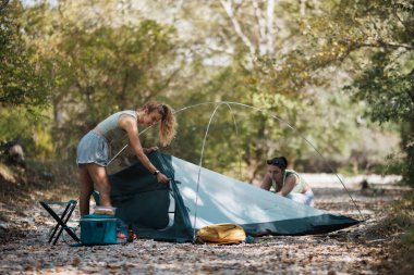 İki arkadaş, ağaçlarla çevrili huzurlu bir nehrin yanında çadırlarını kurmaya hazırlanarak dışarıda dinlenme gününün tadını çıkarıyorlar. Güneş ışıl ışıl parlıyor, neşeli bir atmosfer yaratıyor.