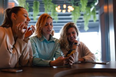 Üç iş kadını şık bir kafede bir sohbete girişiyor, akıllı telefondaki bilgileri incelerken içgörü ve kahkahaları paylaşıyorlar, canlı bir işbirliği ortamı yaratıyorlar..