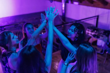 Neşeli bir an, bir grup arkadaşın ellerini havaya kaldırarak modern bir kapalı mekan ortamında dinamik mavi ve mor ışıklar altında birlikte vakit geçirmelerini ve eğlenmelerini gösteriyor..