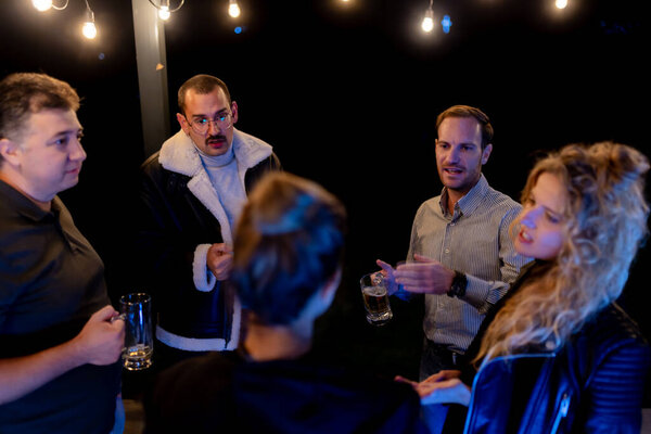 Smiling friends having a casual and cozy gathering at night. Holding drinks and engaging in conversations, illuminated by warm string lights in an outdoor setting.