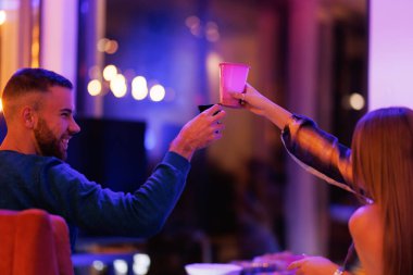 Dostlar arasında paylaşılan canlı bir an. Sıcak kapalı bir toplantı sırasında içkilerle kadeh kaldıran, neşeli bağlantılar gösteren ve gecenin yumuşak parıltısında canlı bir atmosfer..