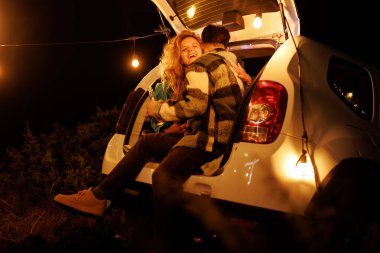 İp ışıklarıyla aydınlatılmış bir arabanın arkasında oturan bir çift kahkaha ve neşeyi paylaşıyor. Çevreleri doğayla çevrili, sıcak ve samimi bir atmosfer yaratıyorlar..